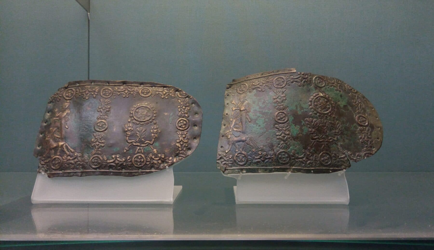 Two corroded Urartian bronze belt fragments on museum mounts, each embossed with banded borders, rosette discs, and small figural scenes.