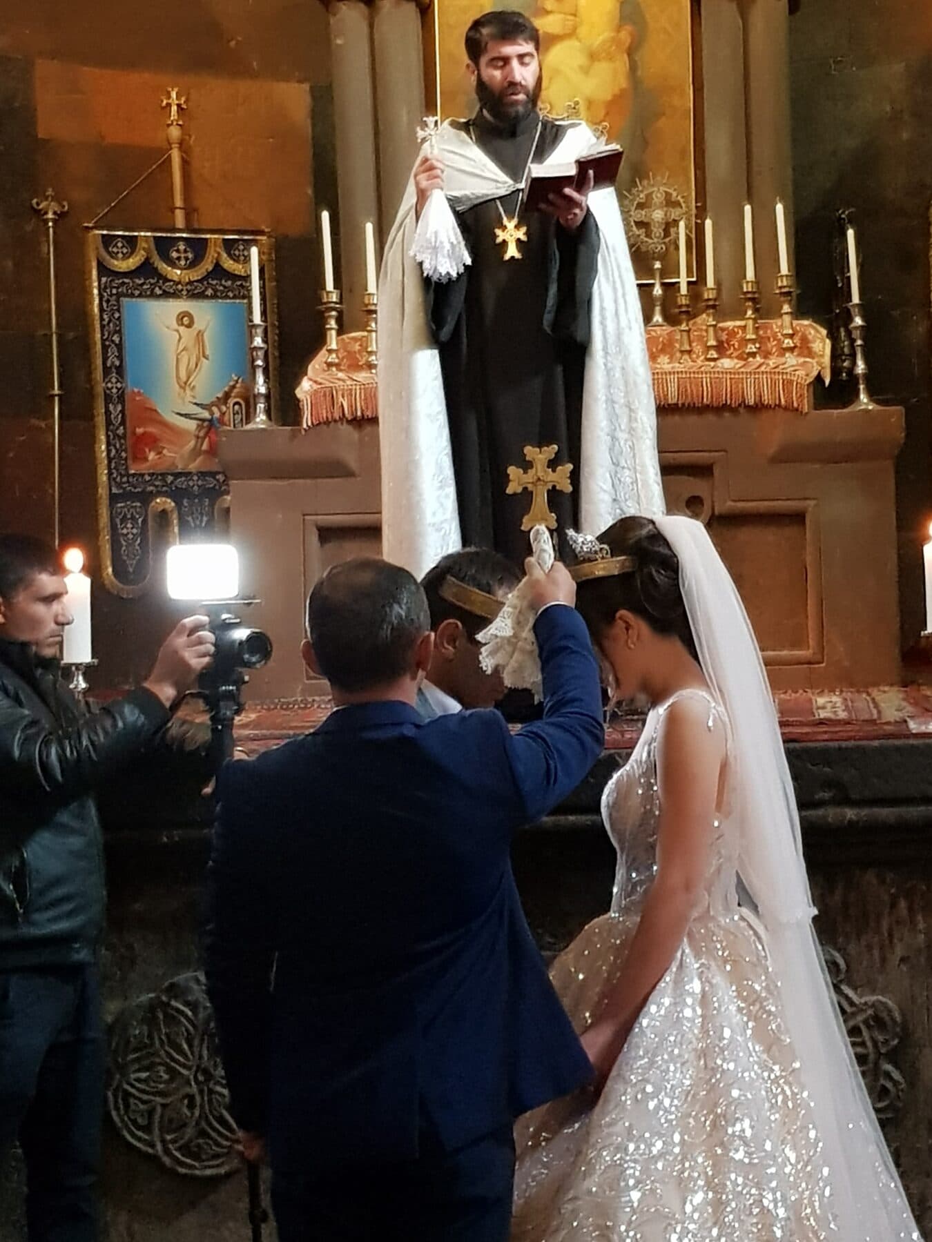 A priest reads from a gospel at an Armenian church altar while a crown is held over the bowed heads of a bride and groom.