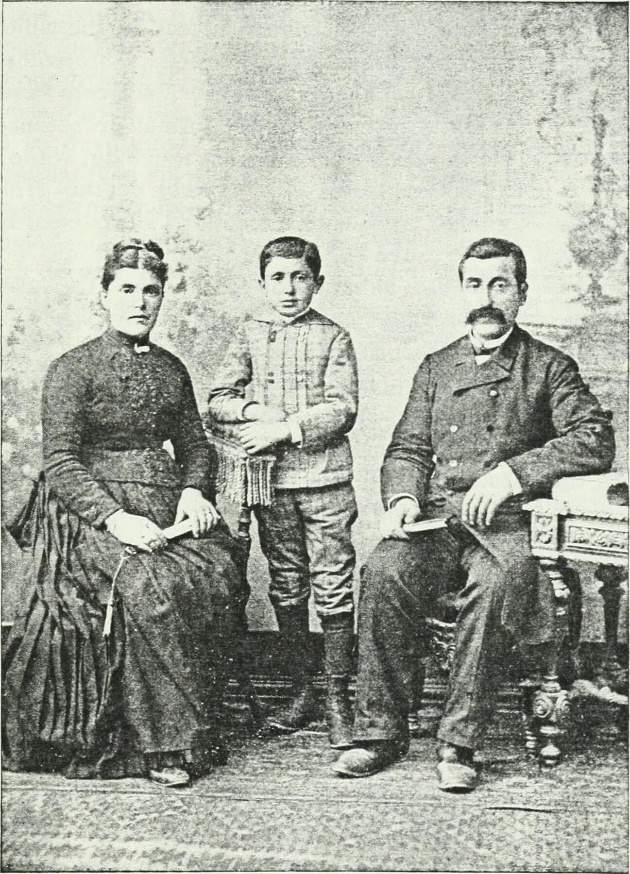 A halftone photograph of an Armenian family of three in nineteenth-century dress: a woman seated at left, a boy standing at the centre, and a moustached man seated at right beside a carved side table.