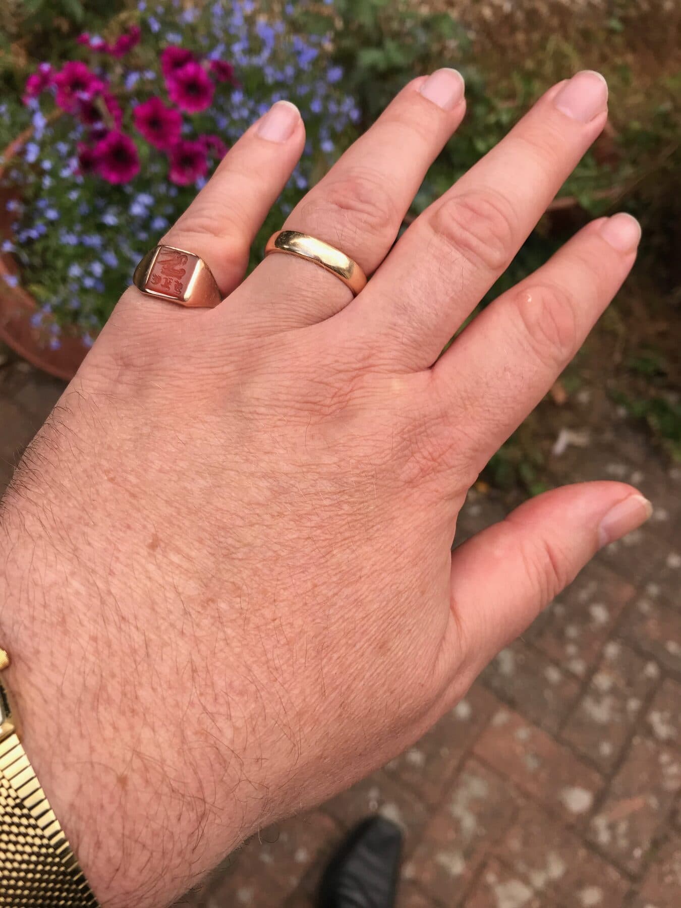 A man's left hand, palm down, wearing a plain gold band on the ring finger and an engraved signet ring on the little finger.