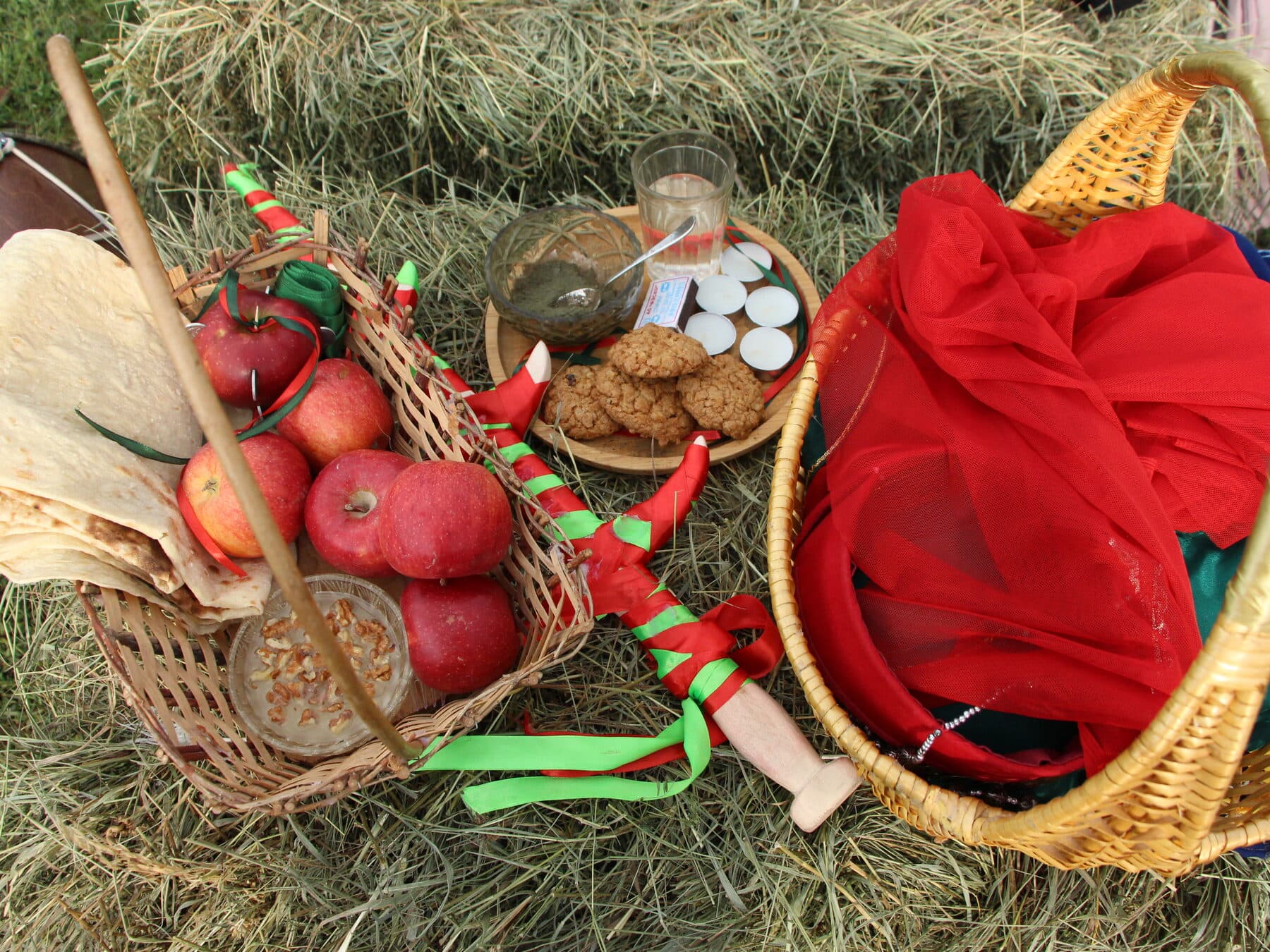 A basket of red apples and lavash bread wound through with red-and-green braided cords, beside a red veil, set on hay among items for an Armenian wedding rite.