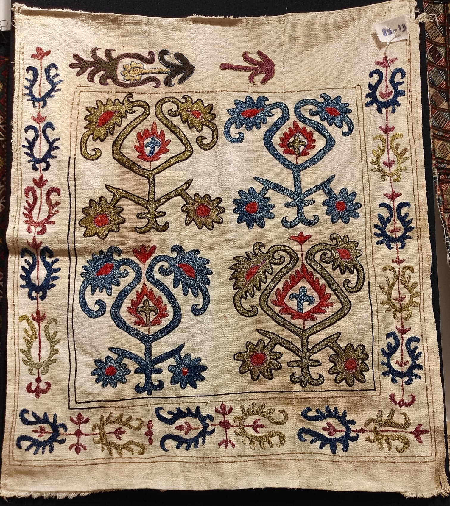 An embroidered cream textile with four symmetrical flowering tree forms rising on central stems, framed by a vegetal border.