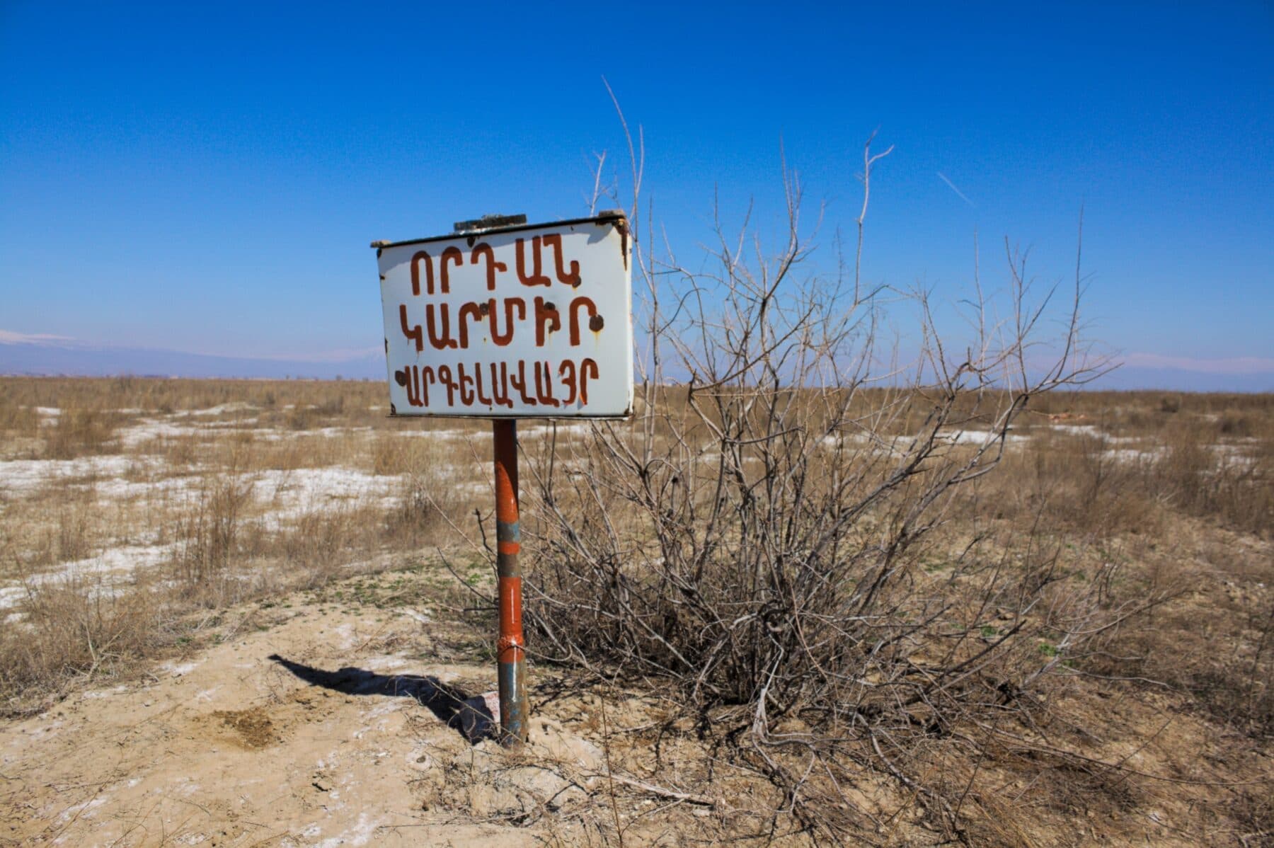 A boundary sign on dry, scrub-covered plain under a clear blue sky, with low hills behind.