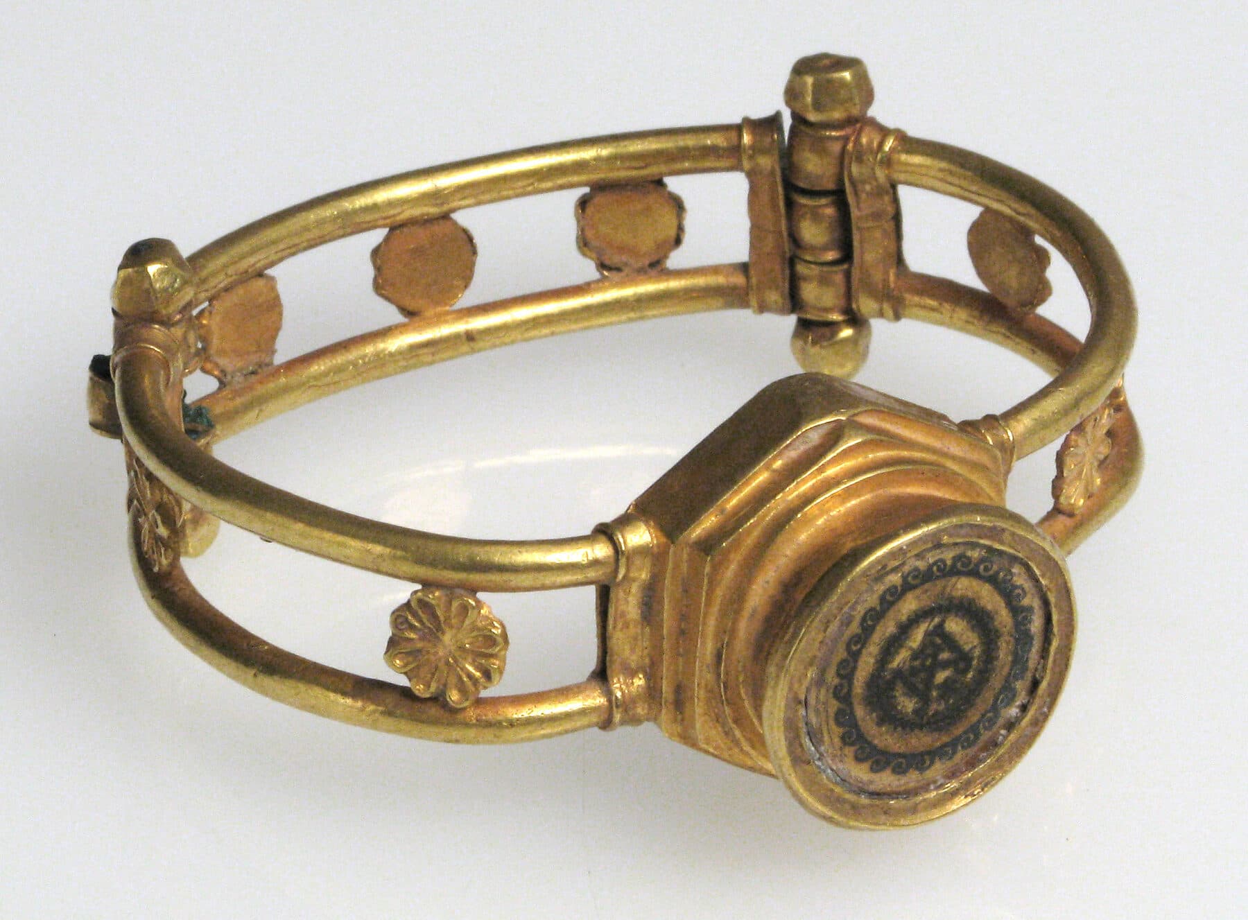 A gold hinged bracelet with an openwork band of rosettes and a raised circular bezel; the bezel medallion is filled with dark niello forming a fine inlaid design.
