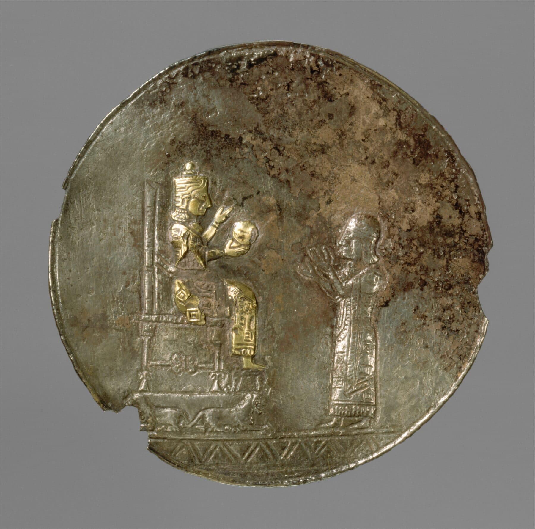 A round silver Urartian medallion with two small gilded figures — an enthroned deity facing a standing worshiper.