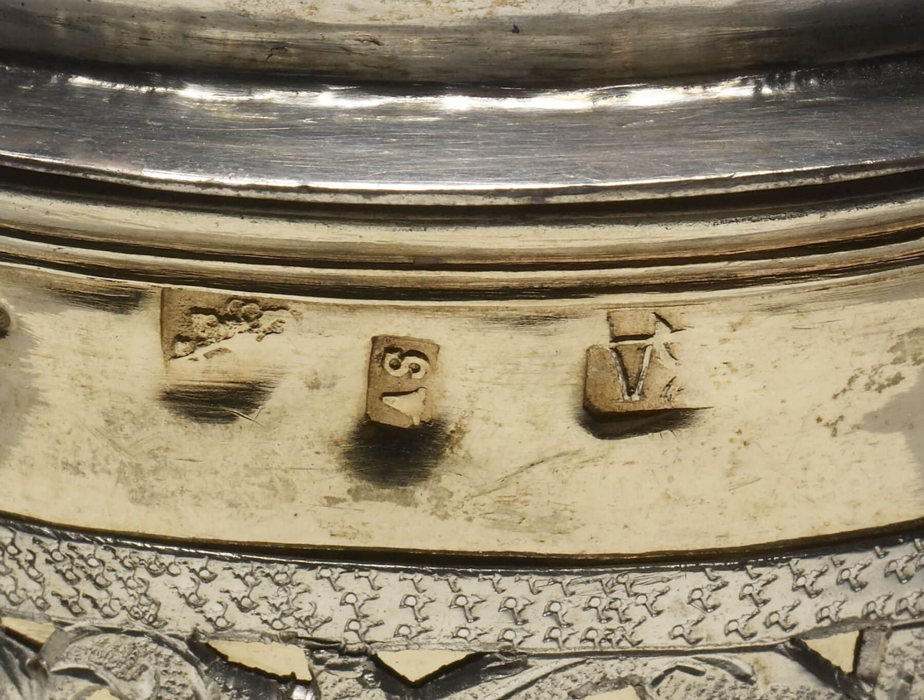 An extreme close-up of a struck mark stamped into the silver surface of a covered cup — a small punched device pressed into the metal.